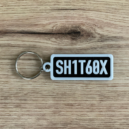An image of the black shitbox keychain with white lettering and border.